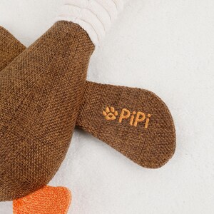Puede incluir: Primer plano de un juguete de peluche marr&oacute;n con una tela texturizada. El juguete tiene una secci&oacute;n acanalada blanca y un acento naranja. El texto "PiPi" est&aacute; bordado en naranja sobre la tela marr&oacute;n. El juguete est&aacute; sobre un fondo blanco.