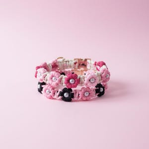 May include: A macrame collar featuring a series of interconnected fabric flowers in shades of pink, white, and black. Each flower center is accented with a small white pearl bead. The collar includes a metallic rose gold-toned buckle closure.