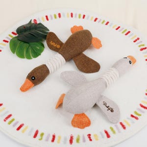 Puede incluir: Dos juguetes de peluche para perros con forma de gansos, uno marr&oacute;n y otro gris, con picos y patas naranjas. Los juguetes est&aacute;n sobre una alfombra circular blanca con un borde colorido y una hoja verde.