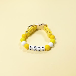 Puede incluir: Un collar de cuentas amarillo con una hebilla metálica y cuentas cuadradas blancas con el nombre LUNA impreso en negro. El collar está construido con cuentas de silicona amarillas redondas e incluye dos cuentas blancas en un diseño circular.