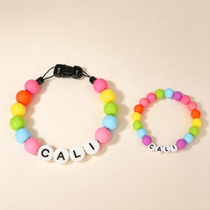 Puede incluir: Un conjunto formado por un collar más grande y una pulsera más pequeña, ambos con cuentas esféricas multicolores y cuentas blancas circulares con el texto negro impreso CALI. El collar incluye una hebilla de plástico de color negro.