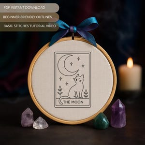 May include: An embroidery hoop featuring a black and white design of a cat, moon, and stars, labeled "THE MOON." The hoop is adorned with a blue and purple ribbon. Amethyst and quartz crystals are placed below the hoop.