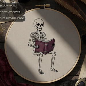 May include: Embroidery hoop featuring a skeleton reading a book with a purple cover. The skeleton is seated, and the design is in black thread on a cream-colored fabric. Text at the top reads "PDF INSTANT DOWNLOAD" and other instructions.