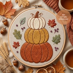 Puede incluir: Un bastidor de bordado con un diseño de tres calabazas apiladas en crema, dorado y naranja, rodeadas de hojas de otoño y bayas bordadas sobre tela beige. El texto PDF PATTERN DOWNLOAD es visible.