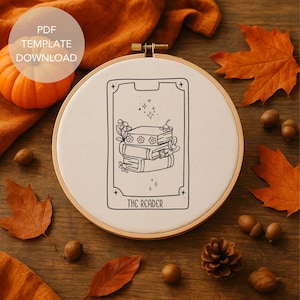 Puede incluir: Un bastidor de bordado redondo con tela crema que presenta un dibujo lineal negro de libros apilados, flores y el texto "THE READER". Hojas de otoño, bellotas y una pequeña calabaza rodean el bastidor.