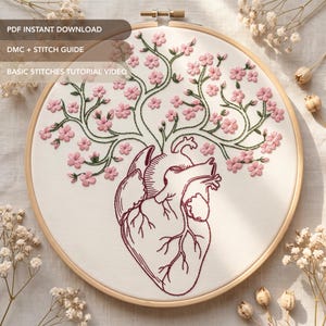 May include: An embroidery hoop containing white fabric with a dark red anatomical heart outline. Green branches extend from the top of the heart, decorated with small pink cherry blossom flowers. The text reads PDF INSTANT DOWNLOAD, DMC + STITCH GUIDE, BASIC STITCHES TUTORIAL VIDEO.