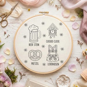 May include: An embroidery hoop with a linen fabric featuring line drawings of a beer stein, cuckoo clock, pretzel, and lederhosen. The words "Beer Stein", "Cuckoo Clock", "Pretzel", and "Lederhosen" are written below each image. The background includes flowers, fabric, and sewing supplies.