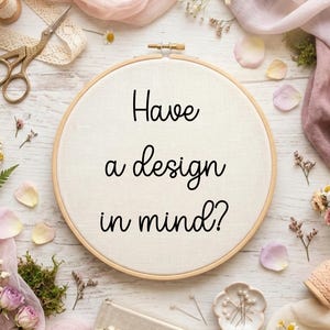 May include: A wooden embroidery hoop with the text "Have a design in mind?" is surrounded by craft supplies. Gold scissors, lace, flowers, and pink fabric are arranged on a light wood surface, creating a craft-themed composition.