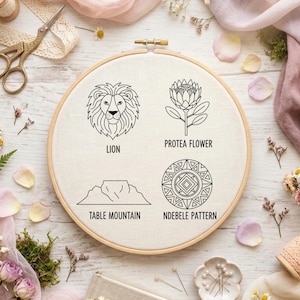 May include: An embroidery hoop with four black line art designs: a lion, a protea flower, Table Mountain, and an Ndebele pattern. The hoop is surrounded by craft supplies, including scissors, thread, and flowers, on a light wood surface.