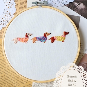 May include: Embroidered dachshunds in colorful sweaters are featured on a white fabric stretched within a wooden embroidery hoop. The dogs are in shades of brown, orange, and yellow, with sweaters in pink, blue, and yellow. The text "Human Made, NO AI" is visible.