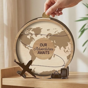 May include: A circular wooden shadow box coin bank featuring a world map background with a dotted travel path. The centre includes the text "OUR ADVENTURES AWAITS" in dark brown lettering. A small aeroplane and luggage icons are at the base.