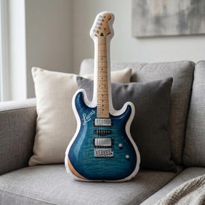 May include: A blue electric guitar-shaped pillow with the name "Lucas" in white script. The pillow has a wooden neck and headstock, and is resting on a gray sofa. The guitar body has a blue finish.