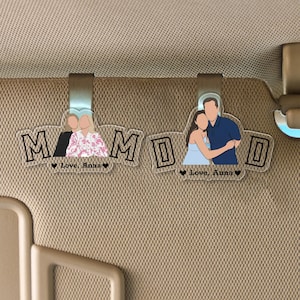 Può includere: Due clip trasparenti per visiera parasole per auto in acrilico con la scritta "MOM" in lettere nere bordate. Ogni clip presenta un'illustrazione personalizzata di persone e il testo "Love, Anna" con un simbolo a cuore.