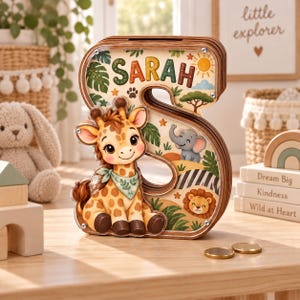 Könnte beinhalten: Eine hölzerne Spardose in Form des Buchstabens S mit einer transparenten Vorderseite. Das Innere zeigt eine Cartoon-Giraffe, einen Elefanten und einen Löwen in einer Dschungelkulisse. Der Name SARAH ist oben in bunten Buchstaben gedruckt. Zwei Goldmünzen.