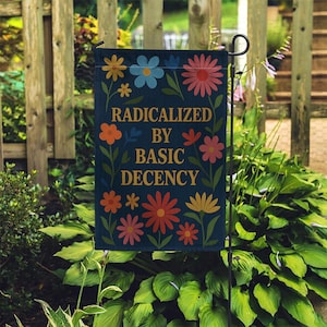 Op de afbeelding: Een marineblauwe tuinvaandel met de tekst "RADICALIZED BY BASIC DECENCY" in gouden letters. De vlag is versierd met kleurrijke bloemen in tinten oranje, roze, geel en blauw. De vlag wordt in een tuin getoond.