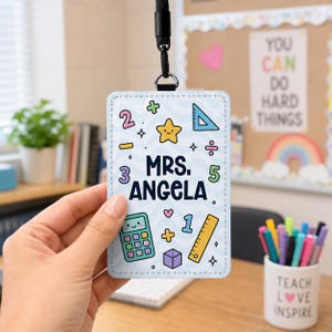 Könnte beinhalten: Ein hellblaues Lehrer-Ausweis-Badge mit dem Namen "MRS. ANGELA" und farbenfrohen Grafiken zum Thema Mathematik. Das Badge wird von einem schwarzen Schlüsselband gehalten. Im Hintergrund ist ein Stifthalter mit der Aufschrift "TEACH LOVE INSPIRE" zu sehen.