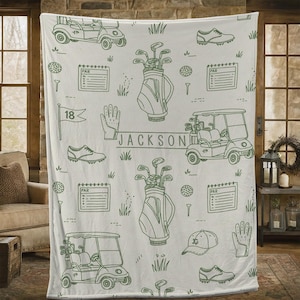 May include: A white blanket featuring a golf theme with green line drawings. The design includes golf carts, golf bags, golf balls, scorecards, golf shoes, gloves, and the name "JACKSON".