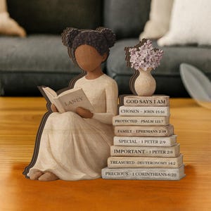 May include: Wooden figurine of a girl reading a book with the name "LANEY" on the cover. A stack of books with religious quotes and a vase with flowers are next to her. The text "GOD SAYS I AM" is on the top book.