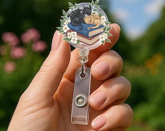 May include: A circular badge reel featuring a graphic of a black dragon and a tan dragon resting on a blue book, surrounded by white flowers and green leaves. The badge reel has a clear plastic strap with a metal snap button for holding an ID card.