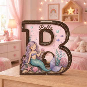 Può includere: Un salvadanaio in legno a forma di lettera B con sfondo rosa, un'illustrazione di sirena, vita marina e il nome Bella stampato in corsivo nero. La lettera ha una cornice marrone scuro, un pannello frontale trasparente e piccole viti metalliche.