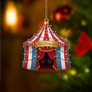 May include: A festive circus tent Christmas decoration, featuring red and white stripes, blue and white vertical stripes, and a red curtain. The ornament is adorned with colourful lights and displays the name "William".