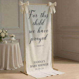 May include: A tall, off-white fabric banner suspended from a gold metal frame with cream-colored bows at the top corners. The banner features black text reading "For this child we have prayed - 1 Samuel 1:27" above "HANNA'S BABY SHOWER 10.10.26".