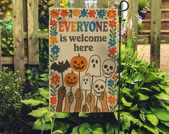 May include: A Halloween garden flag with the text "EVERYONE is welcome here" surrounded by colourful flowers. The flag features illustrations of a bat, pumpkins, ghosts, and skulls held by various skin-toned hands.
