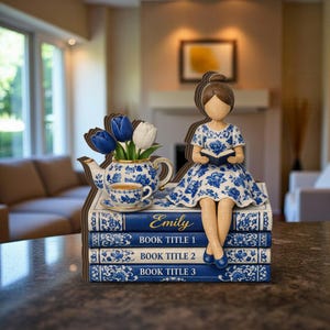 May include: A decorative wooden figurine of a woman reading, sitting atop a stack of blue and white books. A teapot, teacup, and blue and white tulips are on the books. The books are labeled "Emily" and "Book Title 1, 2, 3."