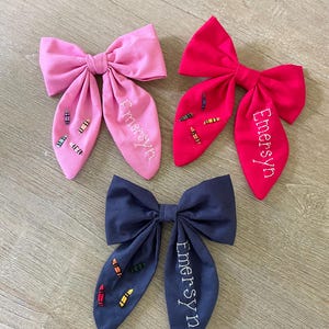 May include: Three fabric hair bows in pink, red, and navy blue. Each bow features embroidered crayons and the name "Emersyn". The bows are arranged on a light wood-grain surface.