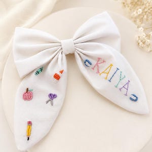May include: A white fabric hair bow featuring colourful embroidered school supply icons including a pencil, scissors, glue, and a crayon, along with the name KAIYA embroidered in multi-coloured thread across the right side of the bow tail.