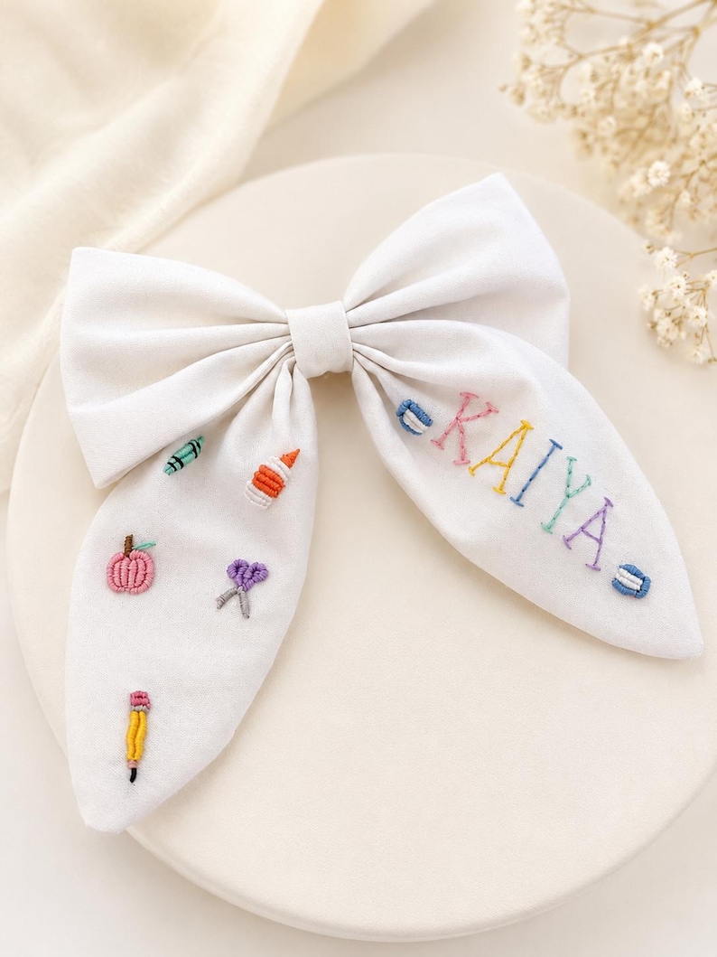 May include: A white fabric hair bow featuring colourful embroidered school supply icons including a pencil, scissors, glue, and a crayon, along with the name KAIYA embroidered in multi-coloured thread across the right side of the bow tail.