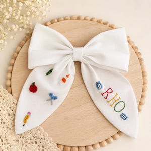 May include: A white fabric hair bow featuring embroidered school-themed motifs including a red apple, blue scissors, a yellow pencil, and a glue bottle. The right side of the bow is embroidered with the name RHO in multi-coloured thread.