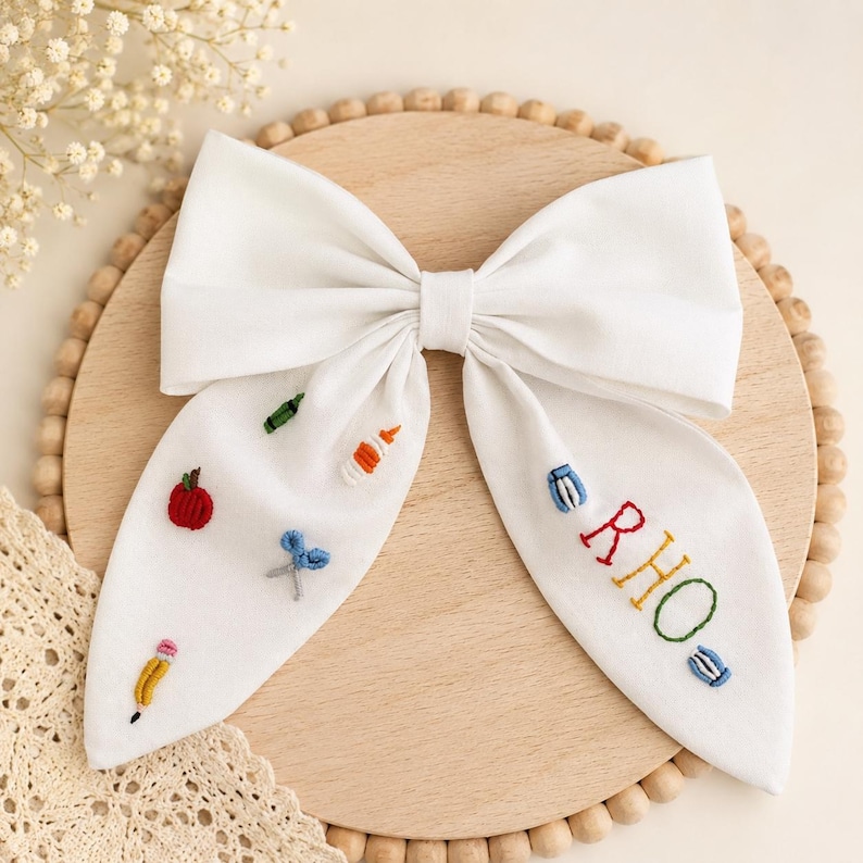 May include: A white fabric hair bow featuring embroidered school-themed motifs including a red apple, blue scissors, a yellow pencil, and a glue bottle. The right side of the bow is embroidered with the name RHO in multi-coloured thread.