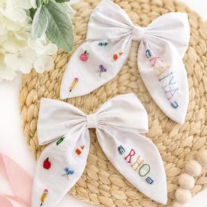 May include: Two white fabric hair bows featuring embroidered school-themed motifs including pencils, scissors, glue, and apples. One bow is personalised with the name KAYA and the other with the name RHO in colourful, multi-coloured stitched lettering.