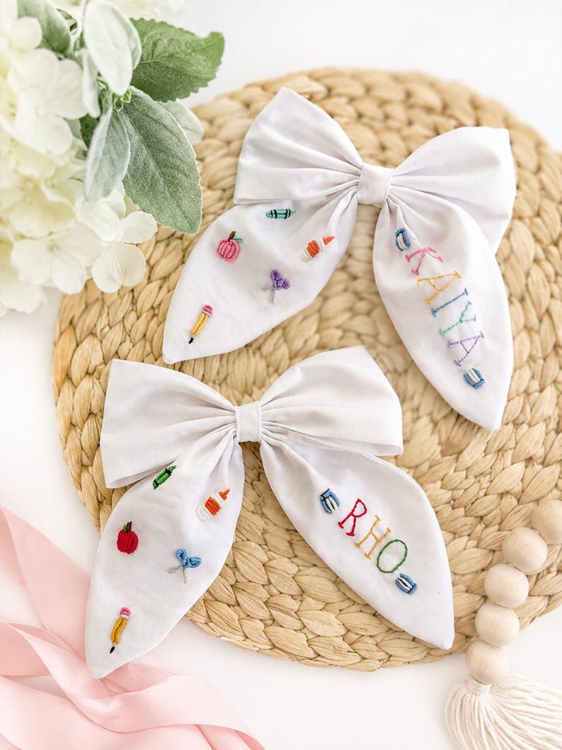 May include: Two white fabric hair bows featuring embroidered school-themed motifs including pencils, scissors, glue, and apples. One bow is personalised with the name KAYA and the other with the name RHO in colourful, multi-coloured stitched lettering.