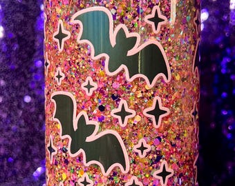 May include: A glitter-filled tumbler with a black bat and star design. The tumbler is covered in pink, purple, and yellow glitter. The top rim is lined with black rhinestones. A fun, festive item for Halloween.