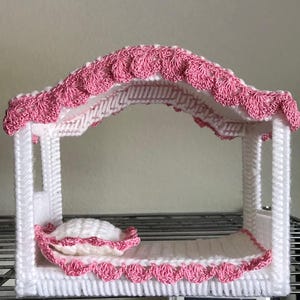 May include: A white plastic doll bed with a pink crocheted canopy and a pink crocheted pillow. The bed is on a metal shelf.