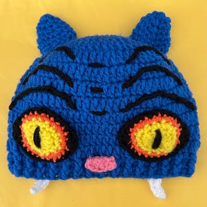 May include: A blue crocheted beanie featuring two pointed ears, black stitched tiger-like stripes, large yellow eyes with orange borders and black pupils, a small pink nose, and two white fangs protruding from the bottom edge of the hat.