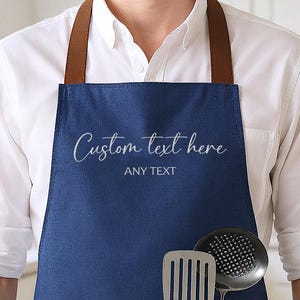 Op de afbeelding: Een marineblauwe schort met bruine leren bandjes en een voorzak. De schort heeft de tekst "Custom text here" en "ANY TEXT" erop gedrukt. Een spatel en een schuimspaan zitten in de zak.