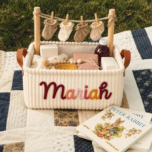 May include: A cream-colored corduroy storage basket with the name Mariah embroidered in red, orange, and pink. It features two wooden posts with a jute string holding four baby socks, brown leather handles, and contains baby items.