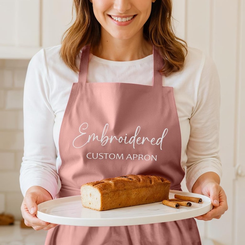 Pu&ograve; includere: Un grembiule rosa antico con la parola "Embroidered" in corsivo e "CUSTOM APRON" a lettere maiuscole. Un pane e bastoncini di cannella sono su un piatto bianco.
