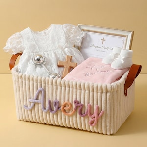 May include: A cream-colored corduroy storage basket with the name Avery in multi-colored yarn lettering. Inside are a white lace dress, a silver rattle, a wooden cross, a pink blanket, white baby booties, and a framed baptism certificate.