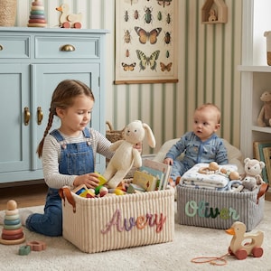 May include: Two rectangular corduroy storage baskets with leather handles. One cream basket features the name Avery in embroidered script, and one gray basket features the name Oliver in embroidered script, both filled with toys.
