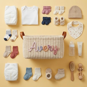 May include: A beige corduroy storage basket with the name Avery in colorful yarn letters, surrounded by baby items including diapers, a white onesie, various knit socks, booties, a beanie, a patterned bib, a bottle, lotion, a brush, and a rattle.