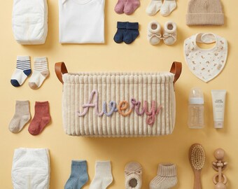 May include: A rectangular beige corduroy storage basket with the name Avery embroidered in multicolored yarn, surrounded by baby essentials including diapers, a white onesie, various knit socks, booties, a beanie, a bib, a bottle, and a brush.