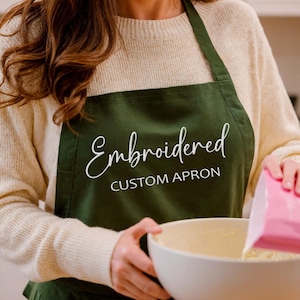 Pu&ograve; includere: Un grembiule verde scuro con la parola "Embroidered" in corsivo bianco e le parole "CUSTOM APRON" in maiuscolo. Il grembiule viene indossato mentre qualcuno versa ingredienti in una ciotola bianca.