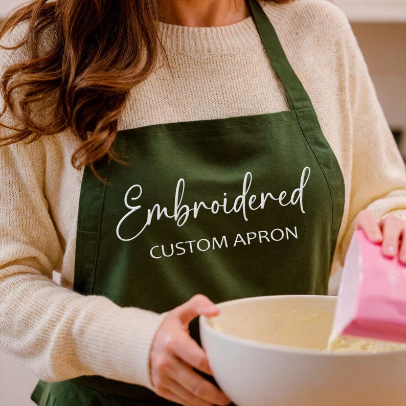 Pu&ograve; includere: Un grembiule verde scuro con la parola "Embroidered" in corsivo bianco e le parole "CUSTOM APRON" in maiuscolo. Il grembiule viene indossato mentre qualcuno versa ingredienti in una ciotola bianca.
