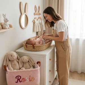May include: A woven wicker changing basket with brown leather handles sits on a white dresser. A pink corduroy storage basket with the name Rosie embroidered on the front holds two beige plush rabbit toys. A white rabbit lamp is on a shelf.