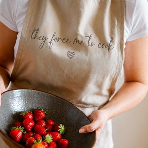 Pu&ograve; includere: Grembiule beige con la scritta "they force me to cook" e un cuore. Una persona tiene in mano una ciotola grigio scuro piena di fragole rosse fresche. Il grembiule &egrave; indossato sopra una maglietta bianca.