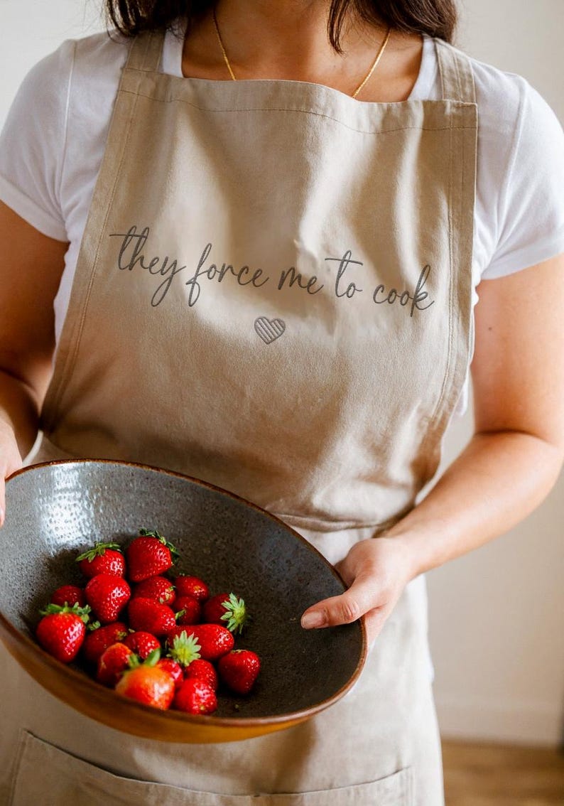 Pu&ograve; includere: Grembiule beige con la scritta "they force me to cook" e un cuore. Una persona tiene in mano una ciotola grigio scuro piena di fragole rosse fresche. Il grembiule &egrave; indossato sopra una maglietta bianca.